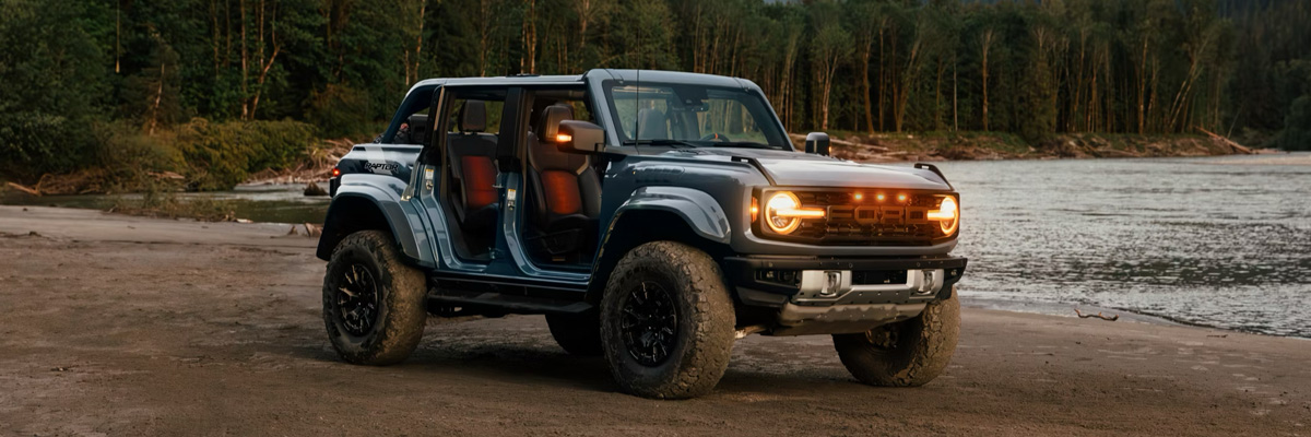 A 2025 Ford Bronco® SUV with the doors off and lights on, parked near a lake in the forest